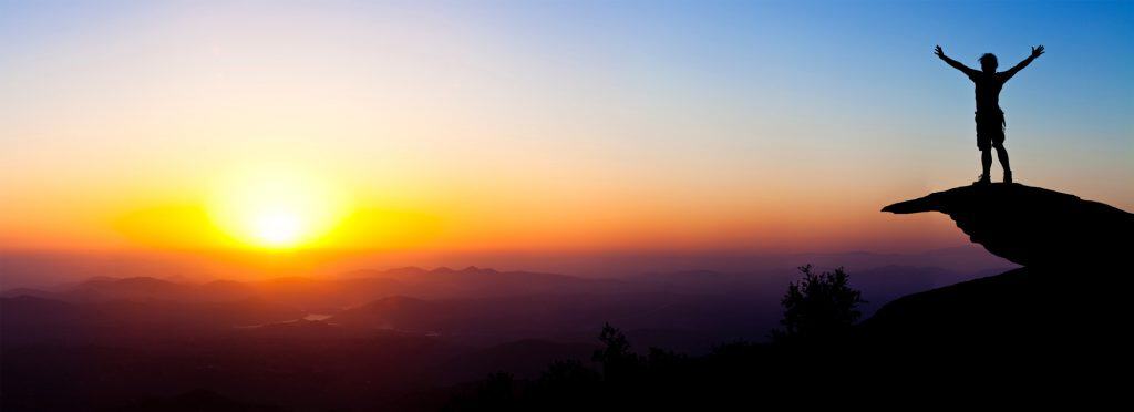 Sunset over mountains with a person standing on a cliff edge, celebrating achievement and freedom.